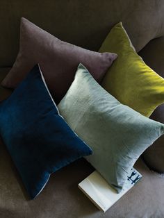 four different colored pillows sitting on top of a brown couch next to a white book