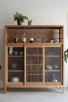 a wooden cabinet with glass doors and plants on top, in front of a white wall