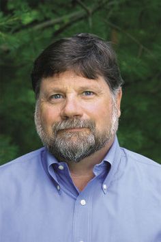 a man with a beard and blue shirt standing in front of some green trees looking at the camera