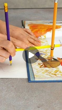 two hands holding pencils and drawing pictures on paper with colored pencils in front of them