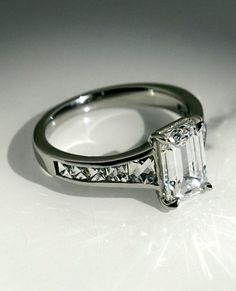 an emerald cut diamond ring with channeled sidestones on a white surface, set in 18k white gold