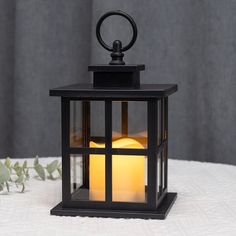 a black lantern with a lit candle inside sitting on a white table cloth next to a plant