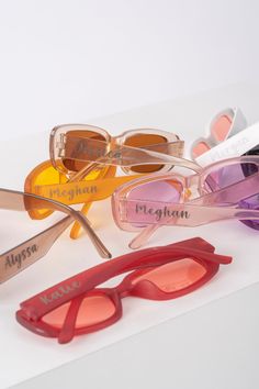 four pairs of sunglasses with names on them sitting on a white surface, one is pink and the other is orange