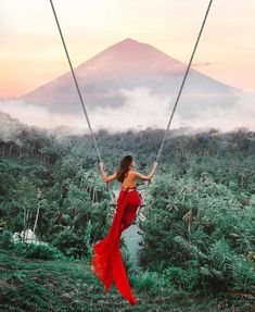 a woman in a red dress is swinging on a rope above the trees and mountains