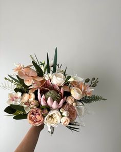 a hand holding a bouquet of flowers against a white wall with greenery and leaves