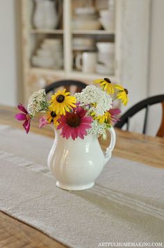 a white pitcher filled with flowers on top of a wooden table next to a dining room table