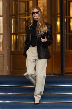 a woman in black jacket and tan pants walking down the street with her hand on her hip