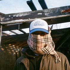 a man wearing a hat and scarf with a plaid bandanna around his head, standing in front of an old wooden structure