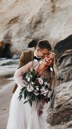 a bride and groom are hugging on the beach with their wedding bouquet in front of them