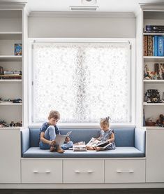 two young children sitting on a blue bench in front of a window with white curtains