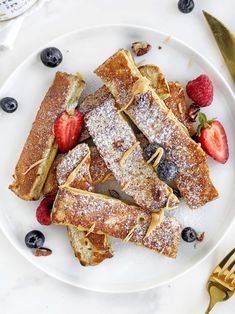 french toast with berries and powdered sugar on a white plate next to gold utensils