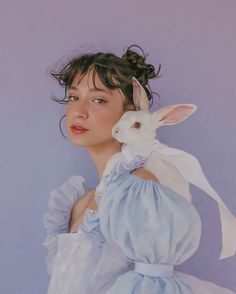 a woman in a blue dress holding a white rabbit on her shoulder and looking at the camera