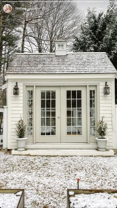 a small white building with two windows and potted plants on the front porch in the snow