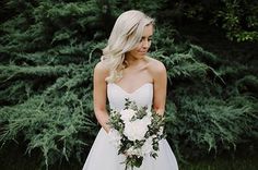 a beautiful blonde woman in a wedding dress holding a bouquet of white flowers and greenery