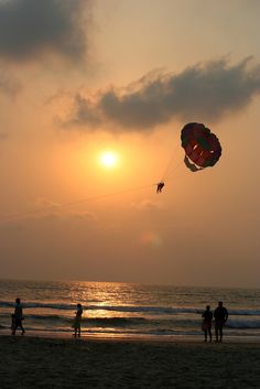 people are flying kites on the beach as the sun sets in the distance behind them