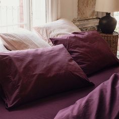 a bed with purple sheets and pillows in front of a window next to a lamp