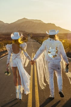 two people dressed in white are walking down the road holding hands and wearing cowboy hats