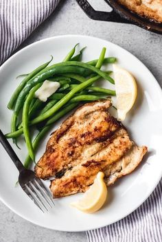 a white plate topped with chicken, green beans and lemon wedges next to a skillet