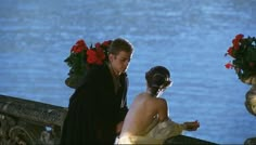 a man and woman standing next to each other in front of flowers on the water