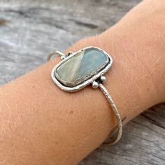 Sterling silver aquateen cuff. Hand made with love! The aquateen has a smooth finish and the silver outline is rather rough, giving the cuff an organic and natural touch. Cuff width: 65 mm (slightly adjustable) Gemstone height: 28mm Gemstone width: 18mm Take a look at our Pebbles & Chance ETSY store https://www.etsy.com/es/shop/pebblesandchance to combine it with other unique, raw, beautiful, precious pieces! Follow us on Instagram! https://www.instagram.com/pebblesandchance/?hl=es One-of-a-kind Bangle Jewelry As Gift, Unique Gemstone Cuff Bracelet As A Gift, Sterling Silver Bangle Bracelet With Natural Stones, Sterling Silver Spiritual Cuff Bracelet With Gemstone, Adjustable Hand Forged Cuff Jewelry, Spiritual Hand Forged Cuff Bracelet Gift, Adjustable Sterling Silver Cuff Bracelet With Natural Stones, Artisan Sterling Silver Bangle As Gift, Unique Sterling Silver Bracelet With Natural Stones