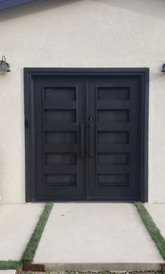 two black double doors on the side of a white building with grass in front of it