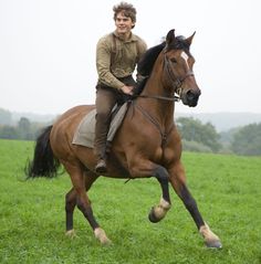 a man riding on the back of a brown horse through a lush green field with trees in the background