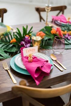 a place setting with pink napkins, goldware and tropical leaves on the table