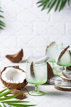 two glasses filled with green liquid next to coconuts and leaves on a white table