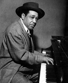 a black and white photo of a man sitting at a piano wearing a suit and hat