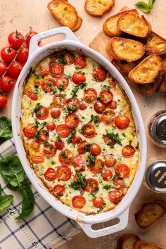 an oval casserole dish with tomatoes and basil on top, surrounded by toasted bread