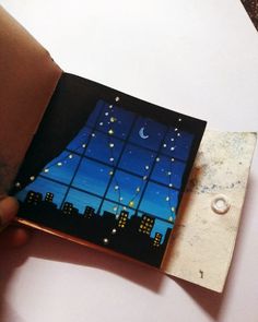 a hand holding an open book with a night scene on the window and stars in the sky