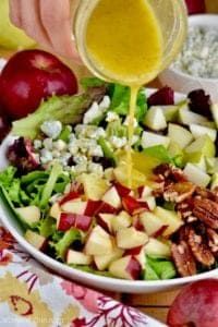 someone pouring dressing into a salad with apples and pecans