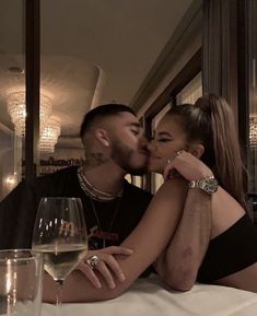 a man and woman kissing while sitting at a table with two glasses of wine in front of them