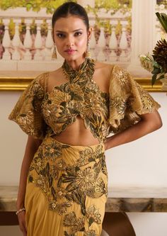 Golden hour is a stunning gold and mustard pre-stitched saree, which features a halter-neck blouse, floral jacket, and a pre-draped skirt. The cut-work adorning the blouse, with olive green floral embroidery, presents sensual glamour, paired up with a pre-draped skirt featuring a modest slit and embellished with floral motifs accentuated with sequins and crystal detailing. Halter-neck blouse adorned with floral embroidery and cut-work detailing as well. Floral jacket accentuated with sparkling beads, crystals, and sequins. Pre-stitched skirt featuring a modest slit embellished with floral motifs and ruch detailing at the backside. Concealed zip on the saree at the left side along with a hook. Blouse contains a single metal zip at the backside. Skirt is connected to the blouse. Halter Neck Blouses, Blouse Designs High Neck, Stitched Saree, Fashion Garments, Floral Lehenga, High Neck Blouse, Hacks Clothes, Unique Blouse Designs, Unique Blouse