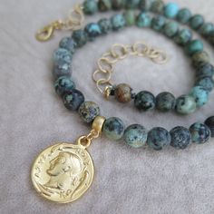 "A gold plated reproduction of a double-sided ancient Greek coin is the striking focal point of this African turquoise beaded necklace. The variegated blue-green coloring of the stones is the reason they are called \"turquoise\", but African turquoise is actually a jasper found in Africa. The gemstones have been finished to a velvety soft, matte patina to add to the necklace's appearance of an ancient and rare archeological find. A gold extender chain completes the necklace and allows it to be a Gold Turquoise Necklace With Gemstone Beads For Spiritual Style, Gold Turquoise Necklace With Round Beads For Gift, Gold Turquoise Necklace With Round Beads As A Gift, Gold Turquoise Necklace With Round Gemstone Beads, Spiritual Gold Turquoise Necklace With Gemstone Beads, Handmade Gold Turquoise Necklace With Round Beads, Gold Turquoise Necklace With Round Beads And Natural Stones, Artisan Turquoise Necklace With Gold Gemstone Beads, Artisan Gold Turquoise Necklace With Gemstone Beads