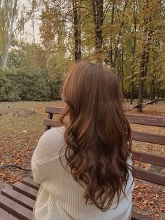 a woman sitting on top of a wooden bench in front of a park filled with trees