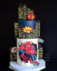 a three tiered cake with spiderman on it's side and city lights in the background