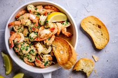 a white bowl filled with shrimp next to bread and lemon wedges on a gray surface