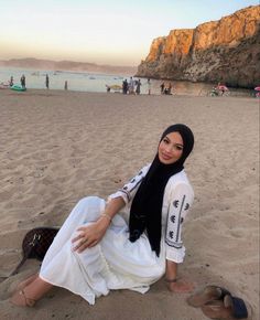 a woman sitting on top of a sandy beach next to the ocean wearing a hijab