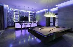 a pool table and bar in a room with purple lighting on the walls, along with two chandeliers hanging from the ceiling