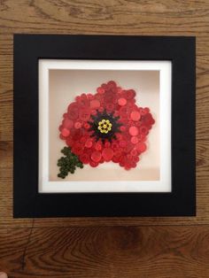 a red and black flower in a white frame on a wooden table next to a person's hand