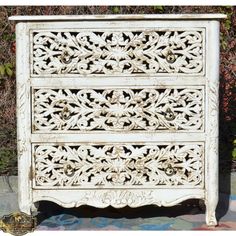 an old white dresser with ornate carvings on the top and bottom drawer, sitting in front of some bushes