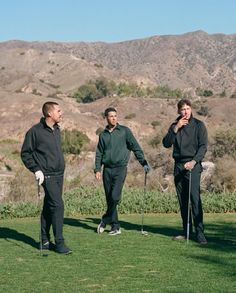 three men standing on top of a grass covered field holding golf clubs and talking to each other