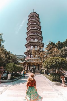 a woman in a dress and hat walking towards a tall pagoda building with trees on both sides