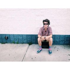 a man sitting on top of a black box next to a white wall and wearing sunglasses