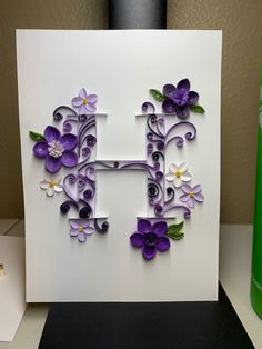 the letter h is made up of paper flowers and leaves on a white card with a green sprayer next to it