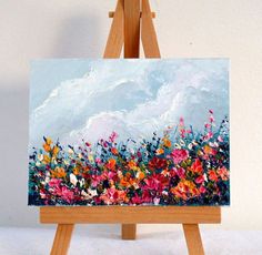 an easel with a painting on it that has flowers in the foreground and clouds in the background