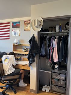 a man sitting at a desk in front of a closet with clothes hanging on it