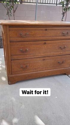 a brown dresser sitting on top of a sidewalk next to a tree and fence with the words wait for it