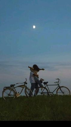 two people standing next to bikes on top of a grass covered field under a full moon